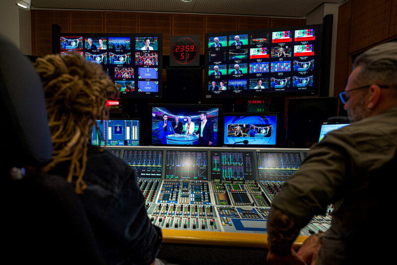 Zwei Personen sitzen in einem Übertragungswagen für´s Fernsehen. Vor ihnen sind viele Monitore, Knöpfe und Schalter.