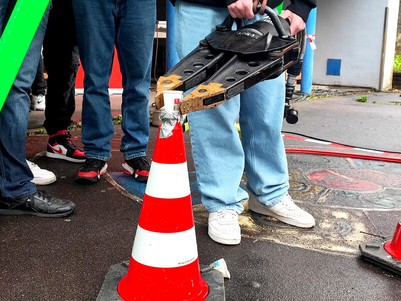 Schüler bei einer Geschicklichkeitsübung mit der Feuerwehr: Mit einer riesigen Zange muss ein voller Wasserbecher von A nach B transportiert werden.