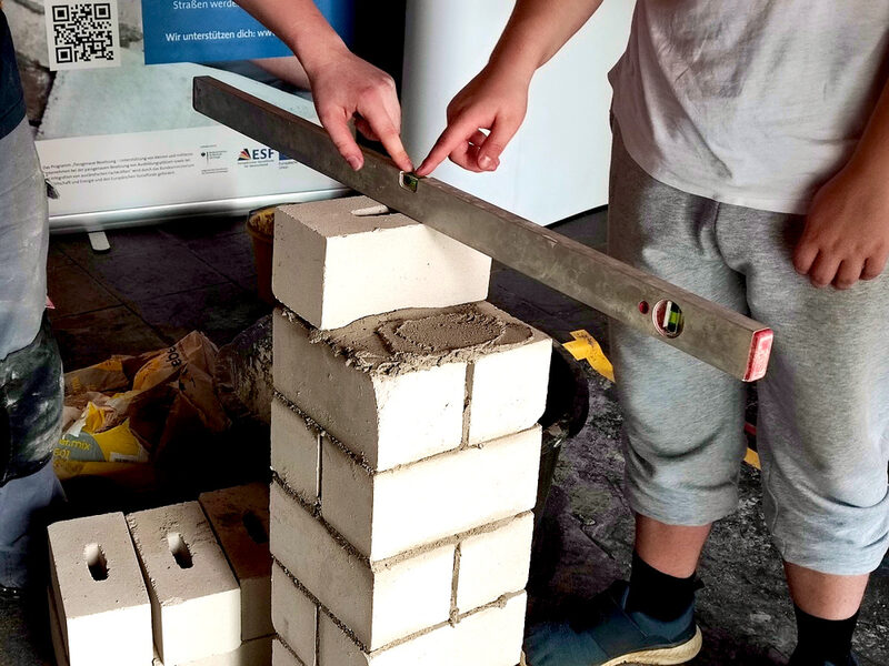 Ein Azubi zeigt einem Schüler, wie man eine gerade Mauer hochzieht.