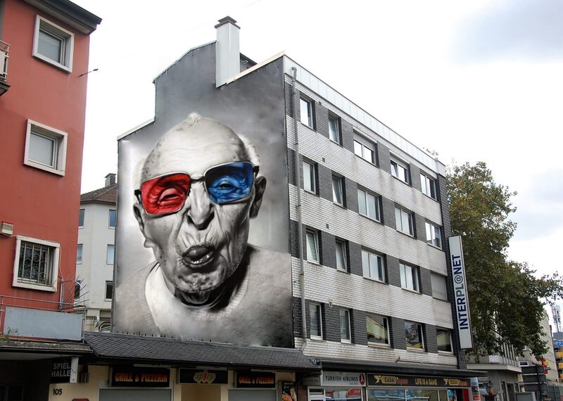 Ein großes Grafiti ziert eine Hauswand. Darauf abgebildet ist eine ältere Person in schwarz-weiß, mit bunter Brille, die die Zunge herausstreckt.
