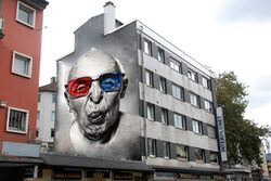 Ein großes Grafiti ziert eine Hauswand. Darauf abgebildet ist eine ältere Person in schwarz-weiß, mit bunter Brille, die die Zunge herausstreckt.