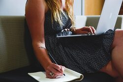 Eine Frau hat einen Laptop auf dem Schoß und notiert sich gleichzeitig etwas.