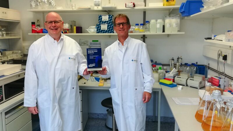 Die beiden Geschäftsführer von AiCuris halten den Rheinland Genial Preis zwischen sich, während sie im Labor stehen.